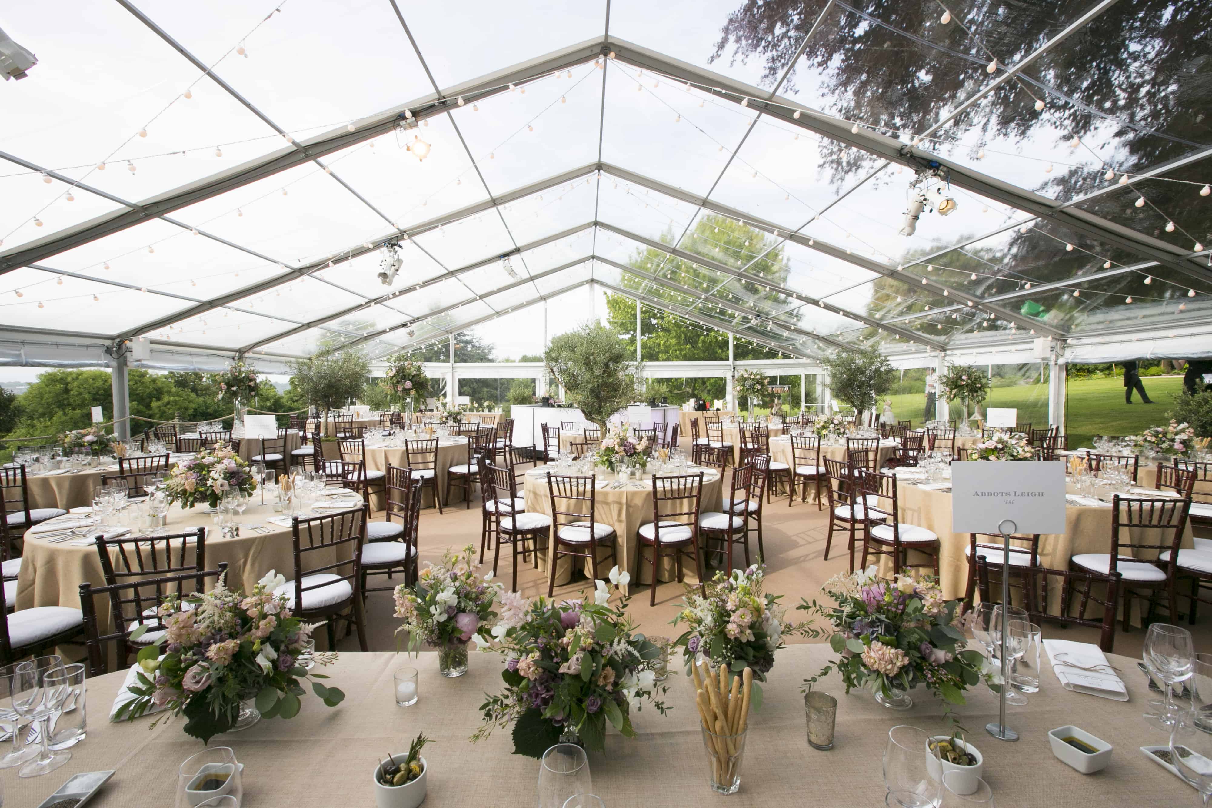 Clear roofed and walled wedding marquee in a countrside setting filled with beautifully dressed tables covered in flowers